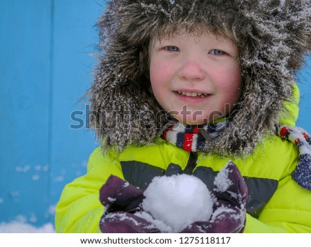Funny boy in bright yellow jacket and big cap with earflaps, playing with snow. Concept of winter sports and children's games. Boy is happy for winter holidays in village.