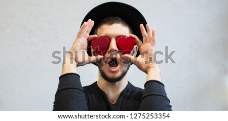 Shocked man holding red hearts on his eyes. Dressed in black, with hat. Valentines day background.