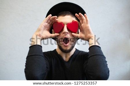 Shocked man holding red hearts on his eyes. Dressed in black, with hat. Valentines day background.