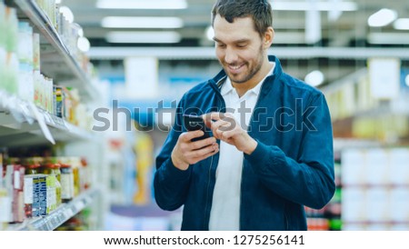 At the Supermarket: Handsome Man Uses Smartphone, Smiles while Standing at the Canned Goods Section.