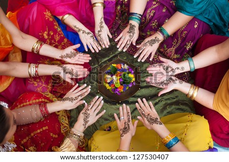 Six pairs of henna decorated female hands arranged in a circle on a colorful background.