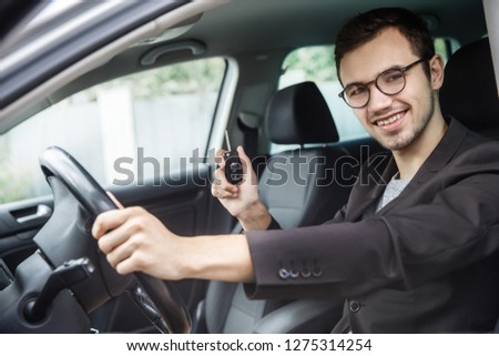 Young man is sitting at his car while looking at the camera. He is holding the keys in his right hand. His left hand is on the steering wheel.