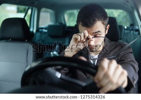 Sleepy young man rubs his eyes with his right hand. His left hand is on the steering wheel. He is sitting at his car. Road safety concept. Royalty-Free Stock Photo #1275314326