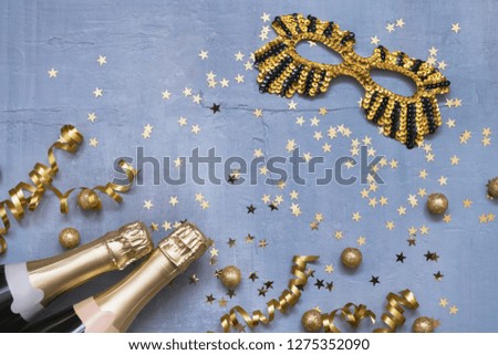 Two champagne bottles with carnival mask, confetti stars and party streamers on blue background. Flat lay of Christmas, anniversary, carnival, New Year celebration concept. Copy space, top view.