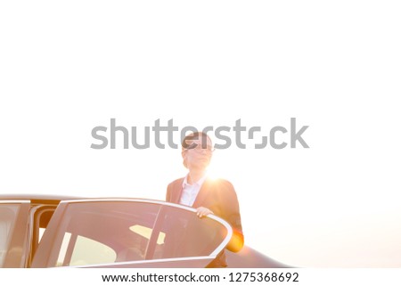 Confident businesswoman disembarking from car against sky on sunny day with a lens flare highlighting the strong sun rays