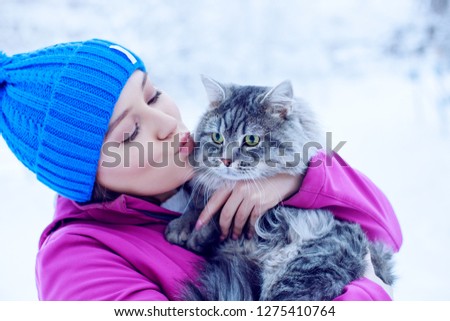 Young woman in winter park kissing her lovely fluffy cat. Girl and her gray cute kitten walking together outdoor. Seasons, pets, friendship, lifestyle concept. Friend of human. Snowy winter day.