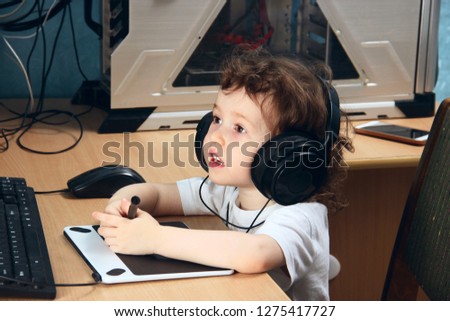 Little 2 3 year old baby girl in white clothers draws at the home computer in graphics drawing tablet.The child is holding a pen and looks at the monitor. Close up.