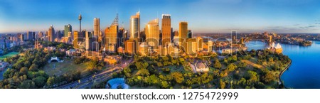 Tall massive high-rise tower buildings of Sydney behind domain and green parklands of botanic gardens on shores of Sydney harbour with major city landmarks in wide aerial panorama.