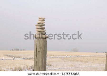 A stack of flat rocks is stacked on top of a wooden fence post, with a cloudy sky and frosty field.
