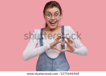 Cute positive European woman makes heart gesture with both hands, expresses love to boyfriend, says be my valentine, wears round spectacles, isolated pink background. Lady shows love sign at camera
