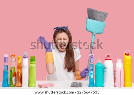 Photo of overjoyed brunette woman keeps hand near face, exclaims and laugh happily, wears headband, rubber gloves, carries broom for sweeping floor, sits against pink background. Time for cleaning
