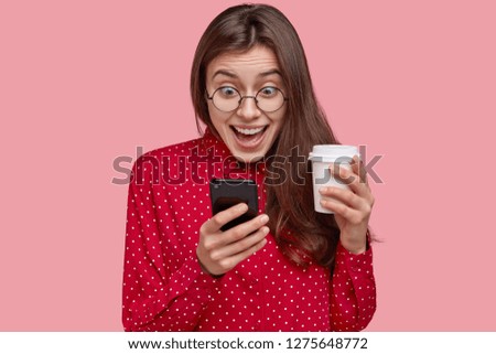 Photo of cheerful young woman checks social networks, plays games on phone, browses online apps, drinks takeaway coffee, dressed in red clothes, isolated over pink background, reads good news