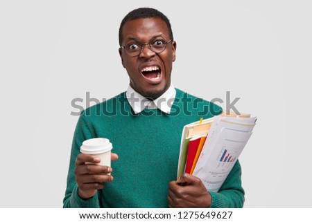 Annoyed dark skinned male frowns face, doesnt want to study economy, market, money and tax, being investment consultant, analyzes company annual report, drinks takeaway coffee, isolated on white wall