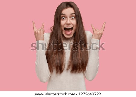 Portrait of emotive dark haired young woman makes rock n roll gesture, listens heavy metal, exclaims loudly, has appealing look, isolated over pink studio wall. Female fun of music likes rock