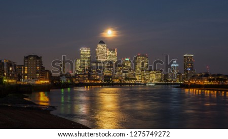 View of Canary Wharf buildings at night under a full moon with light reflections on the water in London, England, United Kingdom