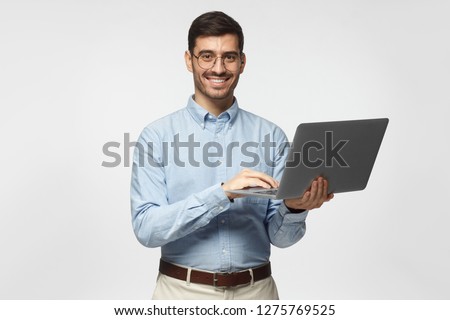 Confident young handsome business man in blue shirt holding laptop and smiling at camera, isolated on gray background
