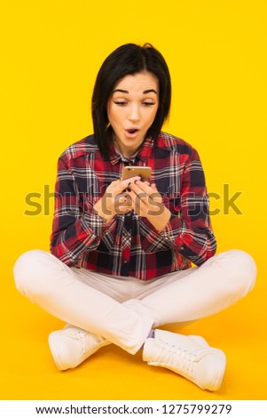 Excited laughing woman in plaid shirt sitting and using mobile phone over yellow background - Image