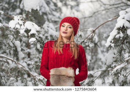 Beautiful woman in red with brown fur cape enjoying the winter scenery in forest. Blonde girl posing under snow-covered trees branches. Young female with snowflakes around in bright cold day, makeup