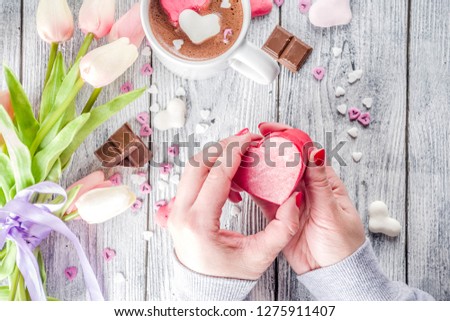 Girl hands hold little heart shaped gift box, old wooden table with hot chocolate with marshmallow hearts and tulip flowers, Valentine day background copy space top view, hands in picture flatlay