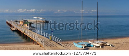 Panoramic photo of the lake shore, pier and sailing boats, with blue sky and blue lake.