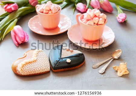 Two pink cups with pink heart shaped marshmallows, pink tulips,  angel figurine and two gingerbreads bride and groom form. Romantic breakfast for two. Close-up, selective focus.