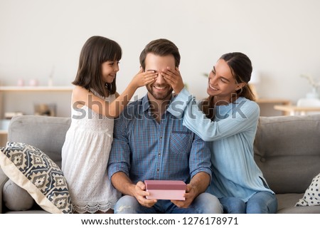 Loving wife and kid daughter making surprise to smiling dad receiving gift on fathers day, family closing eyes of excited daddy preparing present congratulating celebrating happy birthday at home Royalty-Free Stock Photo #1276178971
