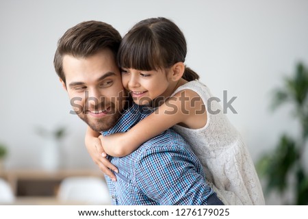 Cute girl embracing dad carrying child on back, little daughter hugging father having fun at home, happy loving daddy giving little smiling kid piggyback ride enjoying good time playing together