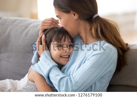 Loving single mother hugging cute little daughter kissing kid sitting on sofa, happy woman mum caressing embracing preschool girl, moms care and support, sincere warm relationships for child concept