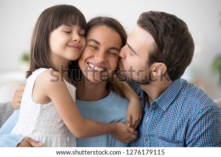 Loving husband and cute kid daughter embracing kissing happy mom wife on cheek congratulating with mothers day, little child girl and dad hugging smiling mum, caring family of three bonding together