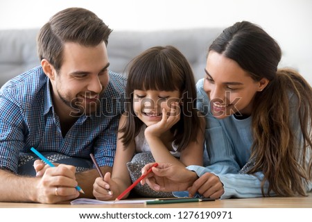 Smiling caring parents and cute little daughter drawing with pencil coloring picture lying on floor, happy friendly family with kid girl play together at home, creative mom dad child weekend activity
