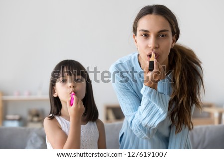 Cute funny kid girl applying lipstick on lips doing makeup imitating copying mom or sister looking at camera, mother teaching child daughter doing make-up together having fun, headshot portrait Royalty-Free Stock Photo #1276191007