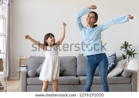 Happy fit sporty mother or baby sitter and funny active child daughter having fun dancing together at home, smiling mom and kid girl enjoy moving to music raising hands standing in living room Royalty-Free Stock Photo #1276191010