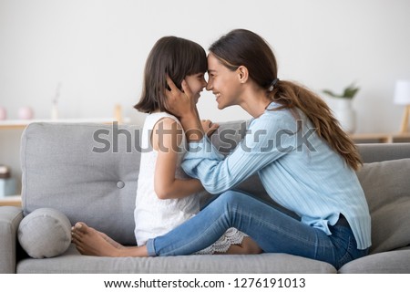 Loving mom laughing caressing touching noses with cute kid daughter sitting on sofa at home, happy mother and little child girl hugging having fun playing enjoy funny moments good time together Royalty-Free Stock Photo #1276191013