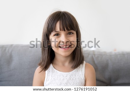 Adorable cute little vlogger looking at webcam, smiling child girl talking to camera making video call vlog communicating online sitting on couch at home, happy preschool kid headshot portrait