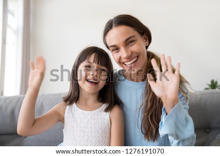Happy mom and cute kid daughter waving hands looking at camera, portrait of friendly mother or baby sitter and child girl smiling talking at webcam saying hello hi or goodbye making online call vlog