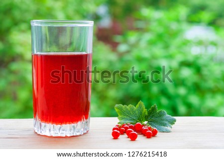 A glass with red juice and branch of redcurrant with leaves on a wooden table on a background of green garden in blur with copy space (shallow depth of field)