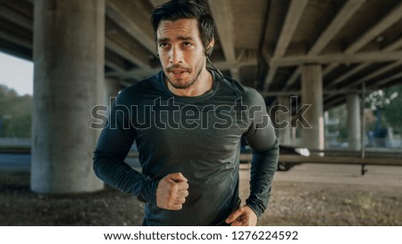 Athletic Young Man in Earphones and Sports Outfit is Jogging in the Street. He is Running in an Urban Environment Under a bridge with Cars in the Background.