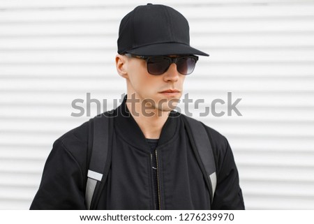 Stylish attractive hipster man in a black basketball cap in a fashionable black sunglasses in a trendy jacket with a black backpack on his shoulders near the metal white wall. American guy.