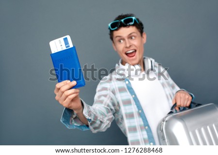 Young guy wearing sunglasses and headphones isolated on gray wall tourism concept standing with baggage suitcase holding passport with plane ticket close-up smiling excited blurred.