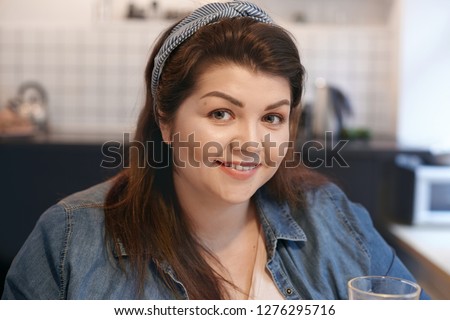 Close up picture of charming beautiful young European woman with chubby cheeks and dark loose hair drinking water or detox fresh juice, choosing healthy lifestyle, smiling broadly at camera