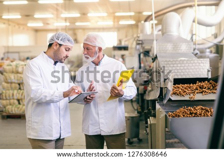 Young Caucasian worker showing to his older colleague statistic on tablet while standing in food factory.