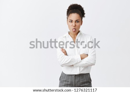 Girl being moody, showing her charachter while having silly argument with girlfriend, holding hands crossed on chest, sticking out tongue and frowning, misbehave and bieng childish over grey wall