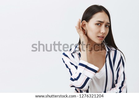 Studio shot of good-looking gloomy girlfriend with long dark hair, leaning towards camera with one ear and gazing at camera, eavesdropping and whispering, hearing rumor, feeling nervous Royalty-Free Stock Photo #1276321204