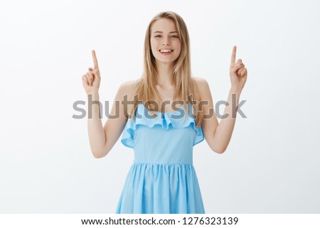 Charming friendly-looking pleasant blonde female in blue dress smiling joyfully at camera as standing with raised fingers and pointing up, showing customers cool product over gray background