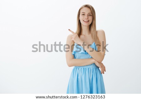 Stylish and confident good-looking woman with fair hair in elegant blue dress asking question as seing interesting thing pointing left and smiling friendly at camera, talking casually over gray wall