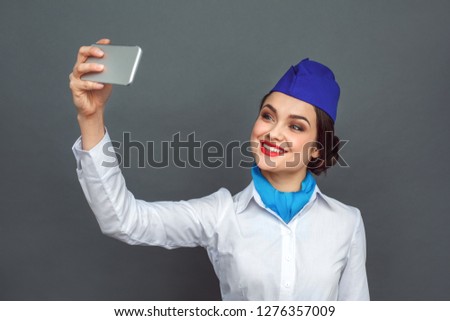 Young woman professional stewardess standing isolated on grey wall taking selfie photo on smartphone looking camera smiling cheerful