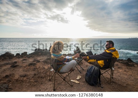 Couple of alternative tourists traveler sit down with the ocean in front and enjoy the rest and the landscape working at laptop connected computer or reading a paper book - sunset and nature 