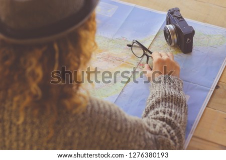 Above and rear view curly woman planning next travel vacation on a paper map - eyeglasses and digital vintage camera on the table - wanderlust and dreamer people 2019 trend