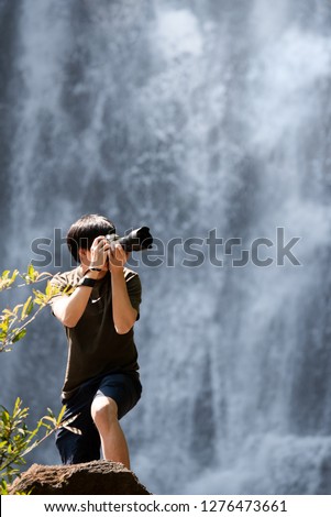Asian man traveler and photographer holding digital camera taking photo at waterfall. Travel lifestyle or nature photography concepts