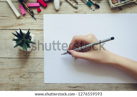 Creative artist workplace flat lay mockup. Top view on table with blank sheet of paper and hand of artist with pencil ready to start sketching or lettering. Art, workshop, drawing concept. Copy space.